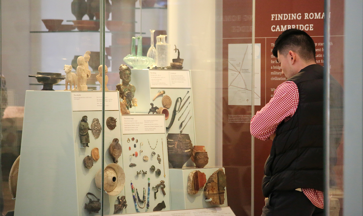 A visitor stands in the local archaeology gallery looking at a case containing Roman finds, including metal figurines, pots, pieces of wall painting and jewellery.