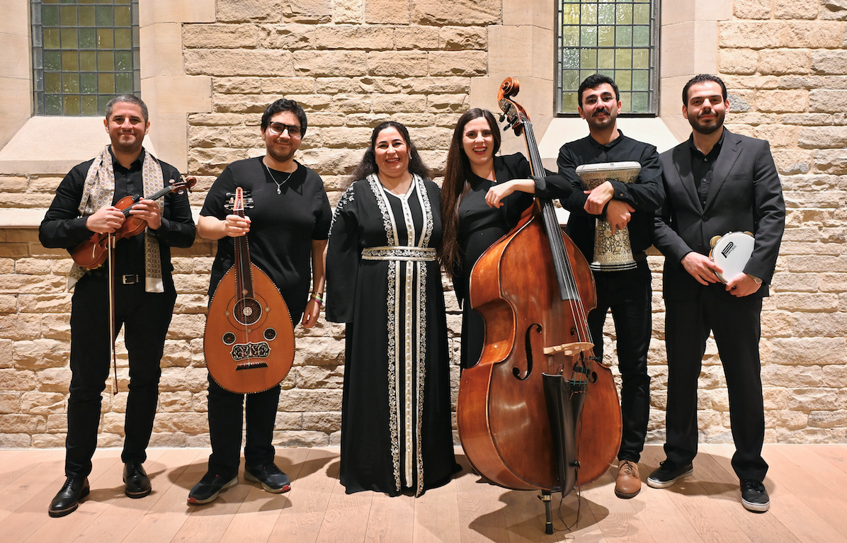 A group of six musicians stand in a line. Two hold percussion instruments whilst three others hold string instruments.