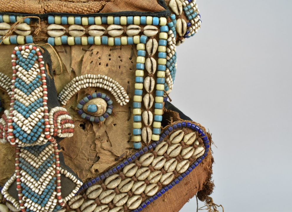 Part of the face of a helmet mask which is decorated with glass beads and cowrie shells. One eye, with its eyebrow, and the nose have all been created through beadwork.
