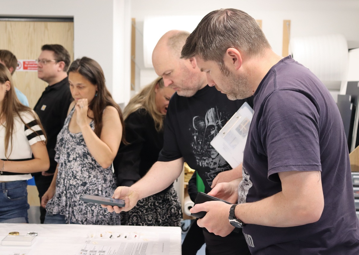 Visitors to the museum's off-site store gather around a table looking down at and photographing objects which have been brought out for them.