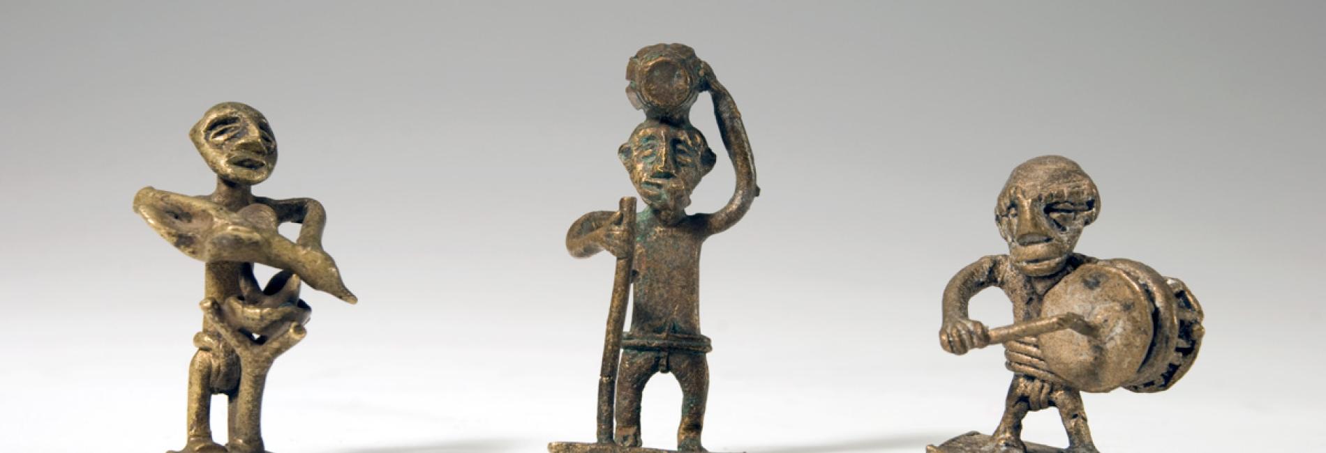 Three Akan goldweights from Ghana, gold in colour and depicting men in various activities, photographed against a grey studio background.