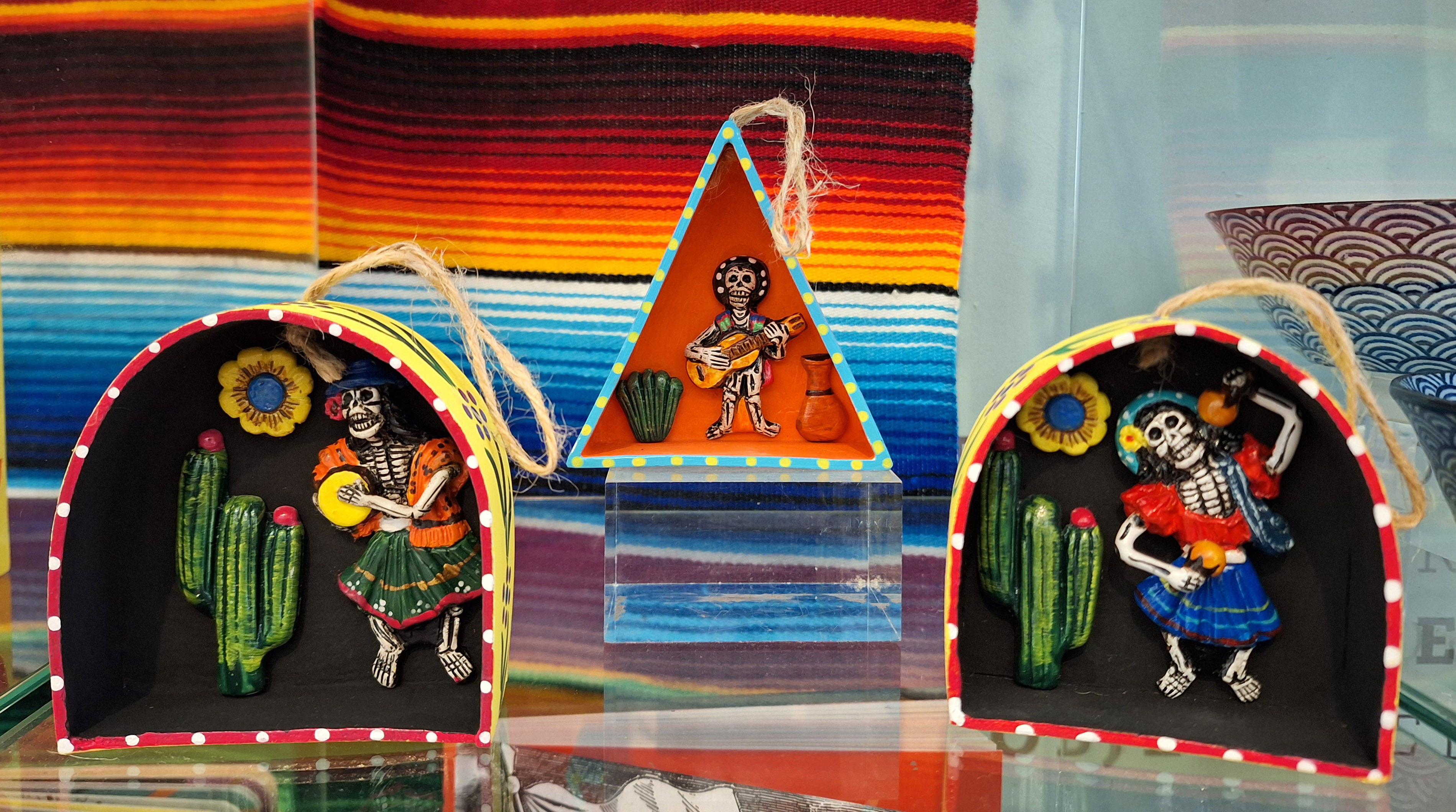 A shop display featuring three retablos. The one in the centre is triangular and is flanked by scenes with square bases and curved tops. Each retablo features a skeleton playing a different musical instrument.