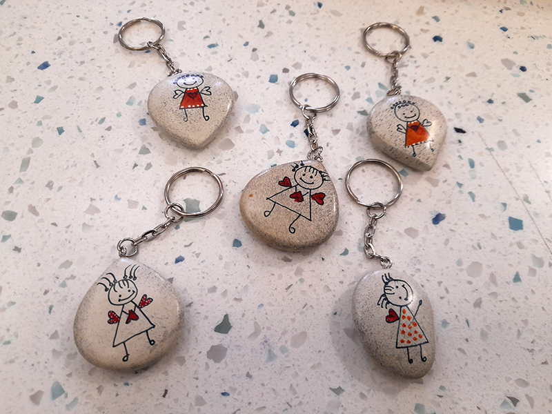 Happy Angel key rings at the Museum of Archaeology and Anthropology shop Five stone key rings with happy angel line drawings on each; angels with red hearts or dresses, each in a slightly different design. Against a terrazzo background of the MAA shop counter.