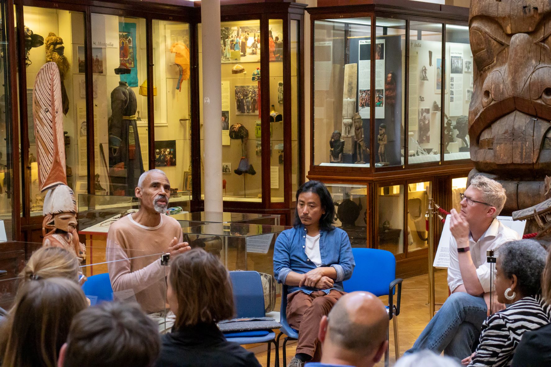 Tony Phillips (left), Temsuyanger Longkumer (centre), and Mark Elliott (right) sit on front of a group of people in the MAA Maudslay Hall. Tony is talking to the group emotively.