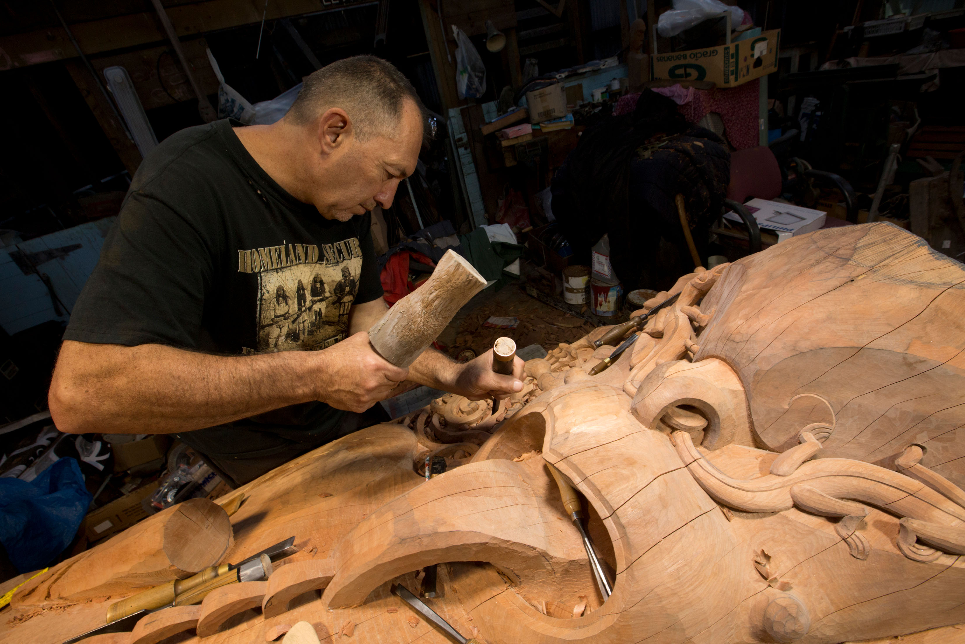 Lyonel Grant carving a large wooden sculpture. He leans over a wooden sculpture with a hammer and chisel, clearly mid work, with a focused expression on his face.