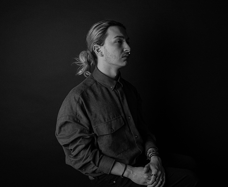 Black and white photograph of a young man with his hair in a bin, sat in half profile with a dark background.