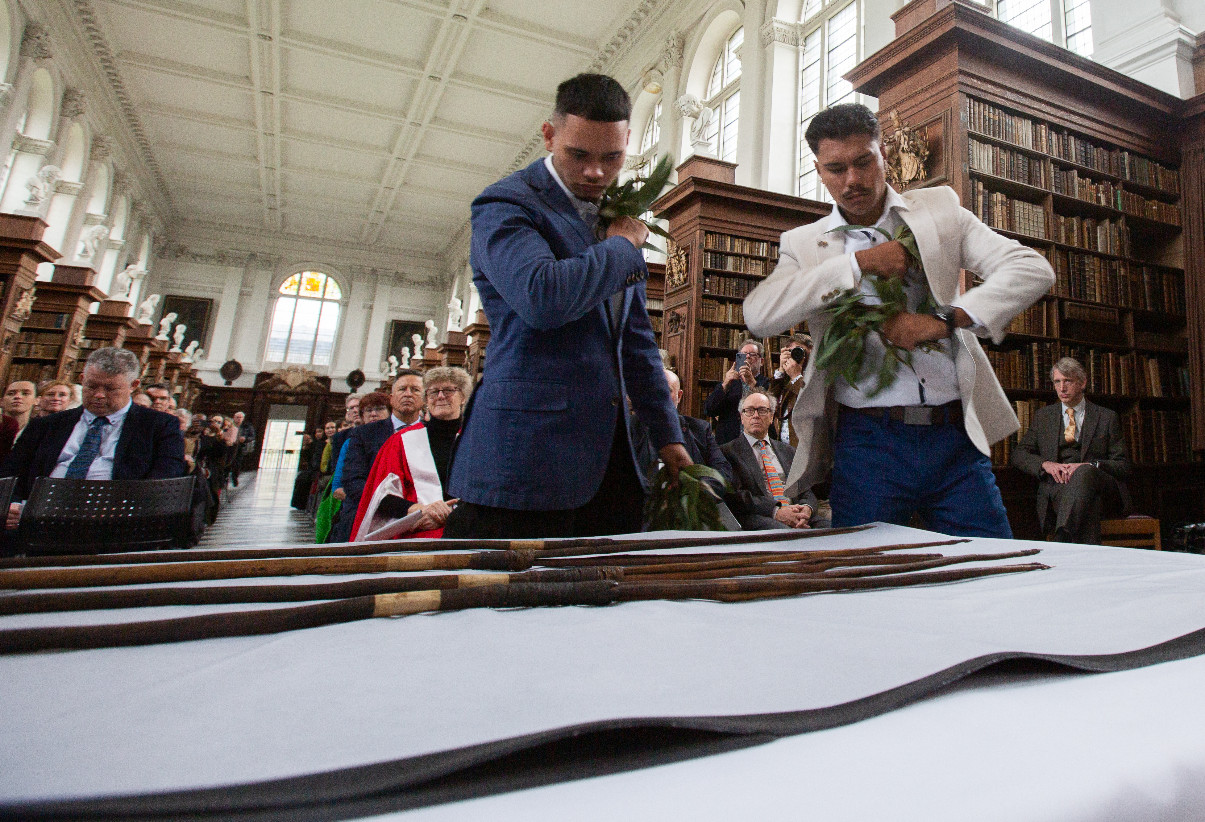Two young men holding leaves in their hands stand over a table with four spears on it. A seated crowd watches from behind them.