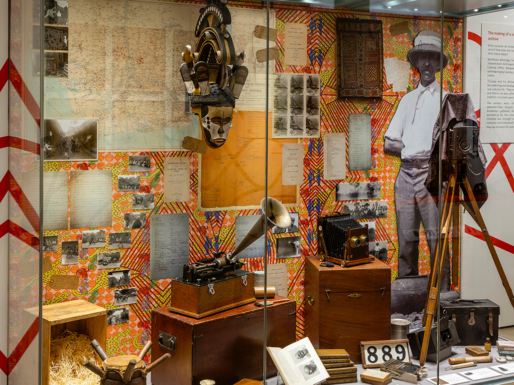 A shot of an exhibition case, with a life-sized photograph of Northcote Thomas, surrounded by some of his possessions used on his tour including a camera. Background is busy and includes contemporary illustration.