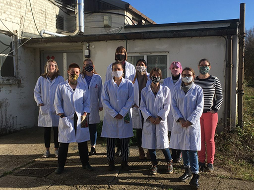 Nine women wearing face masks, eight wearing white lab coats, stand outside a building on a sunny day.