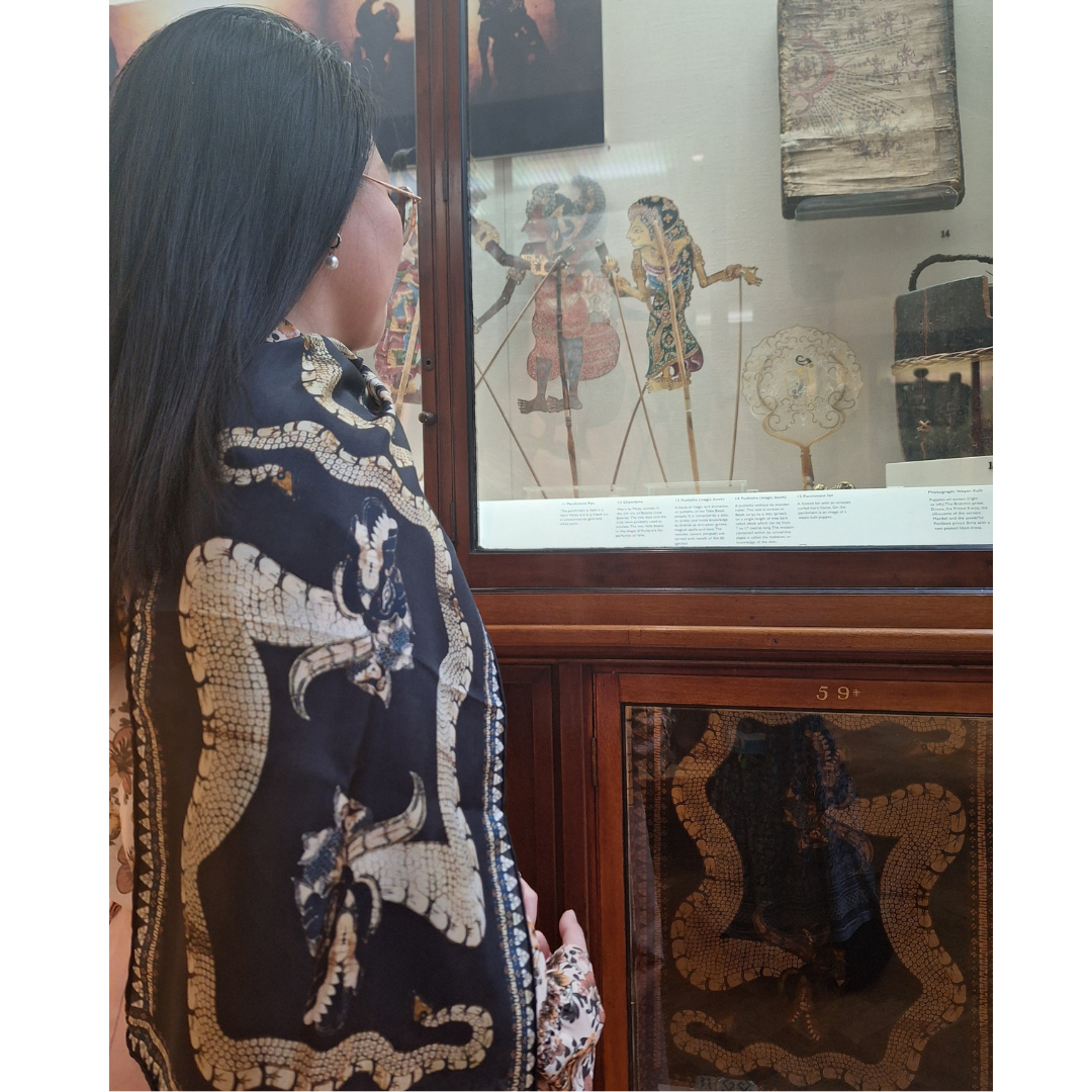 A woman stands in front of a museum display case containing objects from Indonesia, including a batik cloth featuring two dragons. These dragons also appear on the silk scarf she wears draped around her neck and over her shoulder.