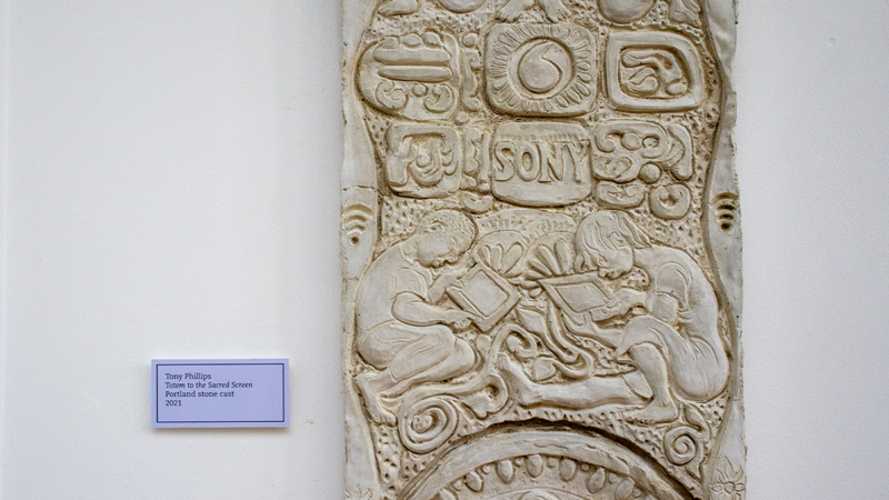 Close up of 'Totem to the Sacred Screen', a carving by Tony Phillips with modern logos (wifi, Sony, and more) and in the centre two children crouching over tablets.