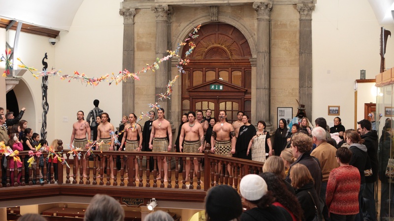 A performance in the museum galleries by a Maori group in front of a packed audience. The women wear black whilst the men are bare chested and wearing a kilt-like garment made from plant fibres.