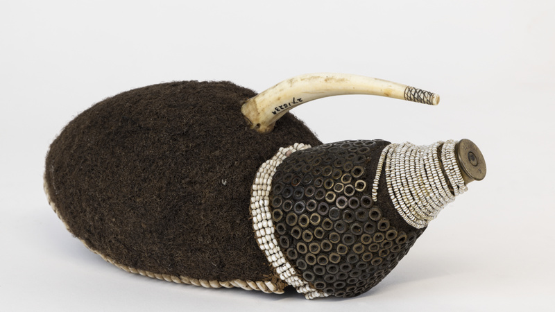 A hollow cap of felted human hair encircled by small pieces of cowrie shell threaded onto a single string. At the front is a curving ivory projection and cone with a foundation of human hair; the base is covered in four rows of annular white beads.