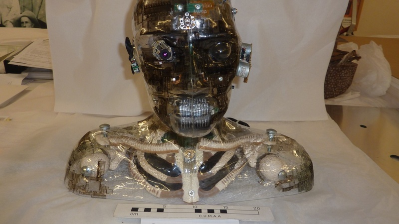 A sculpture of a human head and shoulders, with a digital camera eye, whose observed images are reflected in the screen at the side of the head.