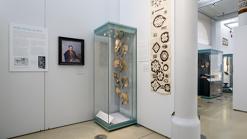 Installation view of the exhibition with barkcloth and fans visible next to a portrait of von Hügel.