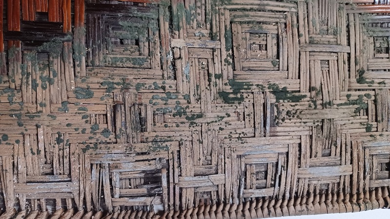 A mat woven with square and cross-shaped patterns. The edge of the mat is discoloured due to water damage and covered in patches of active mould.