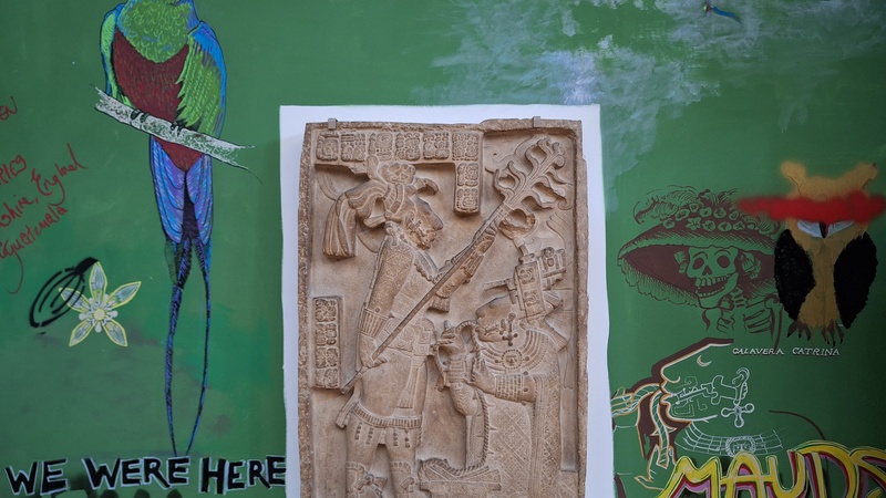 A plaster cast of a Maya carving depicting two figures. The wall around it is painted green onto which graffiti images have been added including a brightly coloured bird, a skeleton wearing a large hat and an owl with a red line over its eyes.