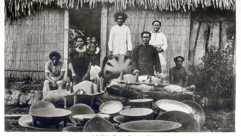 Black and white photograph of men and women standing in front of large bowls, in front of a reed-thatched house.