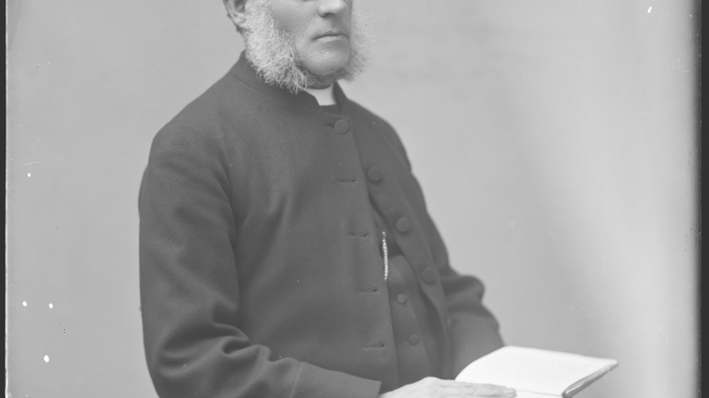 Portrait of a priest, seated, holding a bible. Black and white negative. He has a white beard and wears glasses, as well as a signet ring on his right hand.