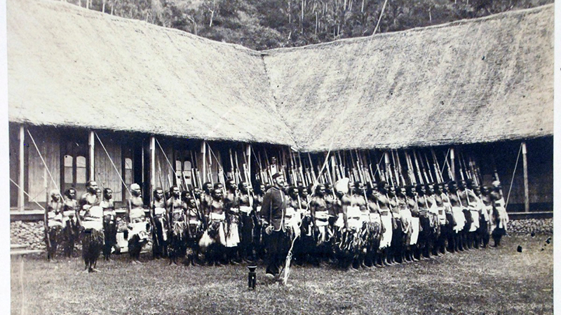 Black and white photograph of a group of people standing in line, with long spears, in front of a house
