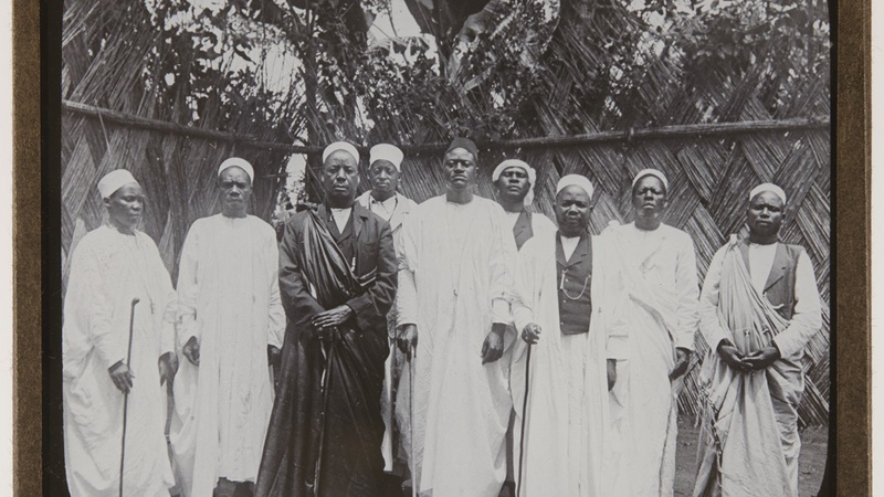 Black and white group portrait of dignitaries and chiefs, mostly in white clothing and one in black. Photograph mounted on a photo slide, with 'Great chiefs of Uganda dressed to attend council. 351' handwritten at the base.