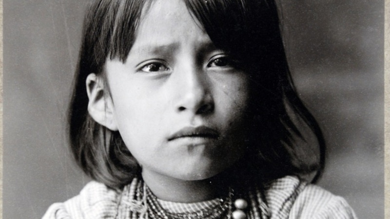 A studio photograph of a Navajo child, with shoulder length dark hair and wearing many necklaces over the top of a checked cotton blouse or dress (uniform).