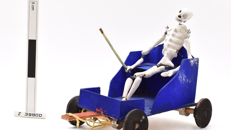  Model of a skeleton sitting in a blue cart. The skeletons joints are made from springs and it is holding a stick. 