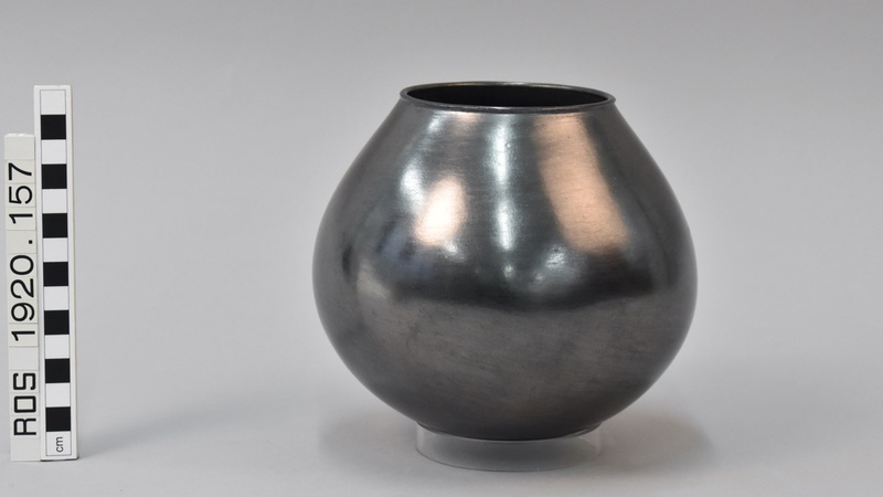 Highly glazed ceramic milk pot, silver in colour, photographed against a studio background. A measure shows it to be around 15cm in height, and shows the accession number 'ROS 1920.157'.