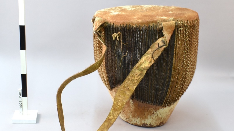 Large conical wooden drum, with a wide top that tapers down to a narrow, truncated base. Both ends of the drum are covered in hide; hair still attached to hide. Sides of drum features closely-spaced lacing made from thin strips of twisted hide, securing b
