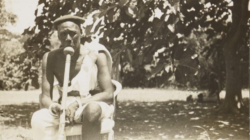 Asepia photograph of an old man, Reth Fatifi Wad Yor, smoking a pipe. He is sat on a chair in front of a large sun-dapped tree scene.