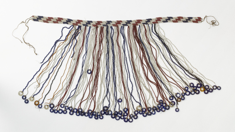 Headband made of a circlet of blue, white and red glass beads in a zigzag pattern. Strings of red, blue, white, black and yellow beads hang down from the circlet, most ending with an annular bead. These are mainly dark blue, but some are clear or yellow.