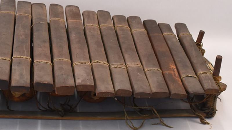 Close up image of the xylophone/balafon, showing that the third key from the right is darker and visibly different than all other keys. The piece is a mid-dark brown wood, with keys tied in with cream coloured string.