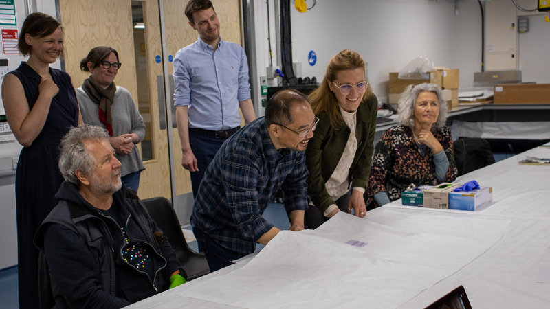 A group of delegates at the Centre for Material Culture, smiling as one man and one woman take part in the activity to handle mystery objects underneath a cloth.