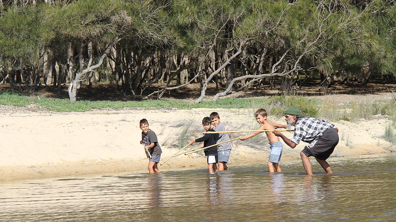 Rod Mason crouching with a spear, showing four young boys how to fish, in shallow waters near a beach.