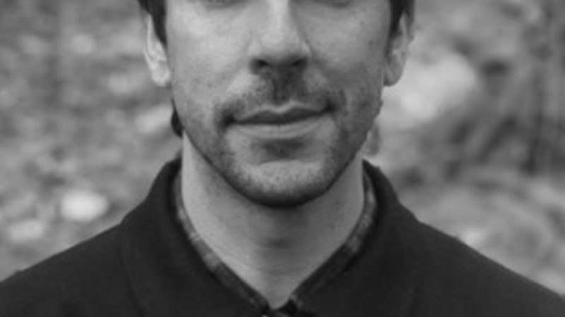 A curator standing outside looks into the camera. The image is black and white and he has short dark hair and stubble. He wears a dark coat.