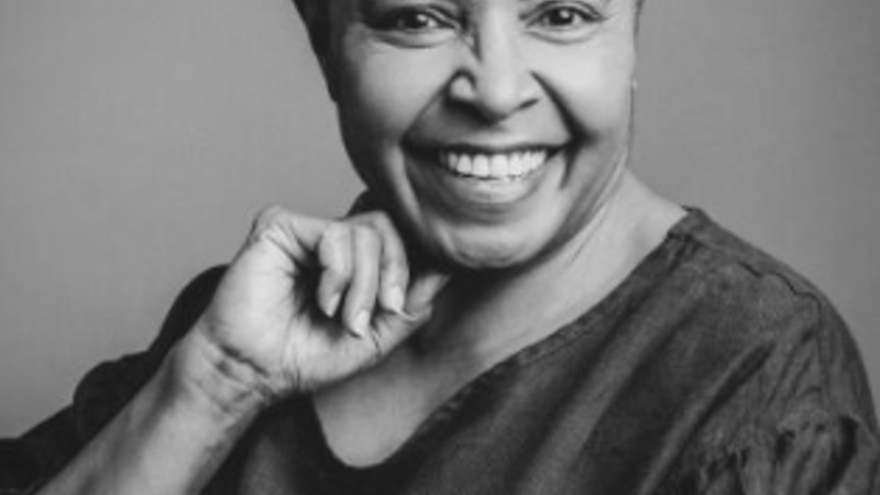 A widely smiling curator looks towards the camera. The image is black and white and she has short dark hair and wears a dark top. Her right handis raised and rests near her jaw.