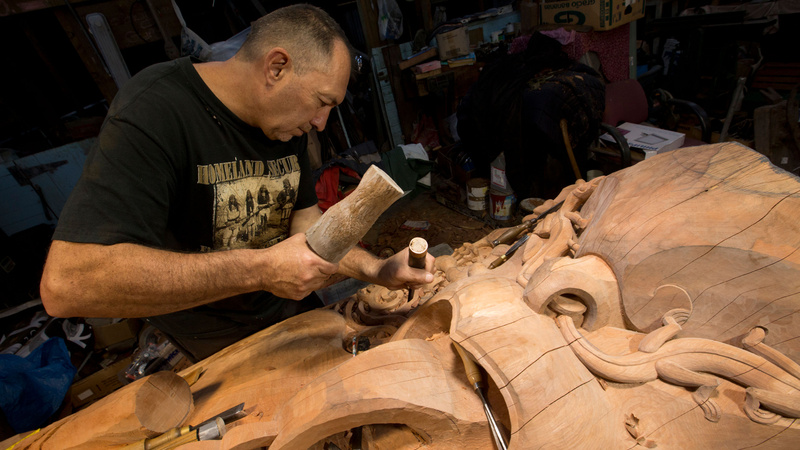 Lyonel Grant carving a large wooden sculpture. He leans over a wooden sculpture with a hammer and chisel, clearly mid work, with a focused expression on his face.