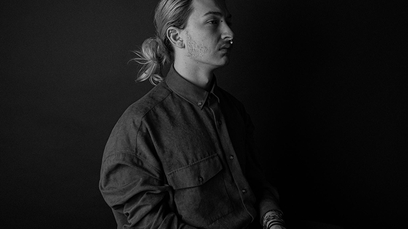 Black and white photograph of a young man with his hair in a bin, sat in half profile with a dark background.