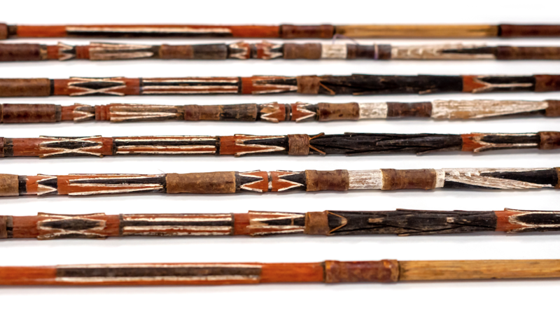 The wooden shafts of a collection of arrows which have been carved and painted using red, white, and black pigments.