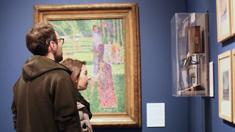 Two visitors to the museum stand in front of a painting. They are looking at objects displayed on the wall adjacent.