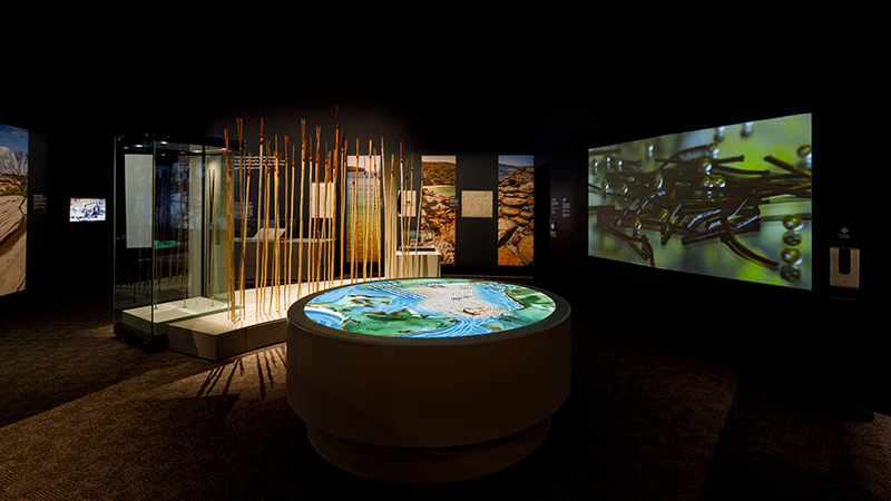 Exhibition installation scene with spears visible before a round map installation, and a video installation. The room is dark.