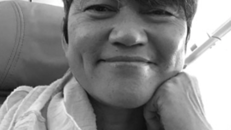 A smiling curator looks towards the camera resting her jaw on her left fist. The image is black and white. She has short and her glasses are push onto the top of her head.