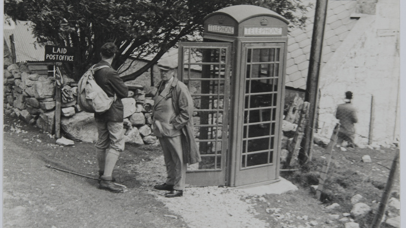 Black and white photo of James Wordie and a second expedition member standing next a British telephone box and a sign pointing to “Laid Post Office”. Village buildings are in the background.