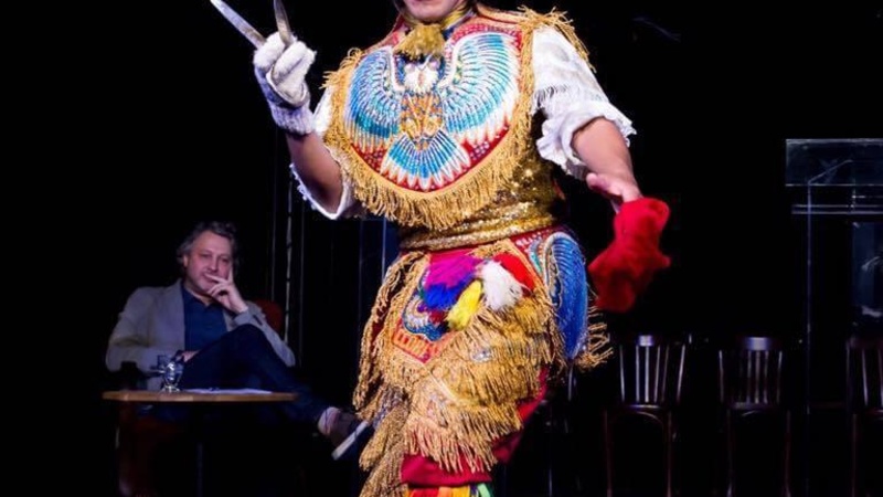 A dancer performs, possibly on a stage. They wear a brightly coloured Peruvian costume and hold a pair of scissors in one hand. There is a large rectangular hat on their head depicting the face of a bird, possibly an owl. There is a full depiction of the 