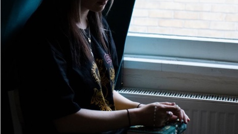 Photograph of a woman sat down, holding her grandfather's possessions, against a window. The lighting is deep and melancholy.