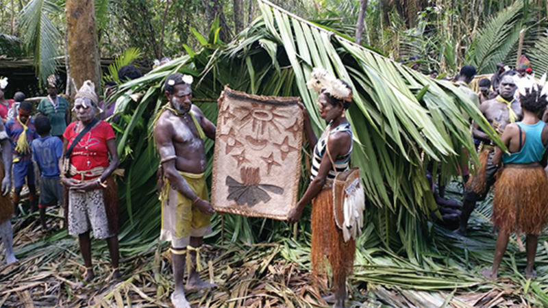 Asmat villagers hold a pit mat in the forest. A man and a woman hold the mat with people surrounding.