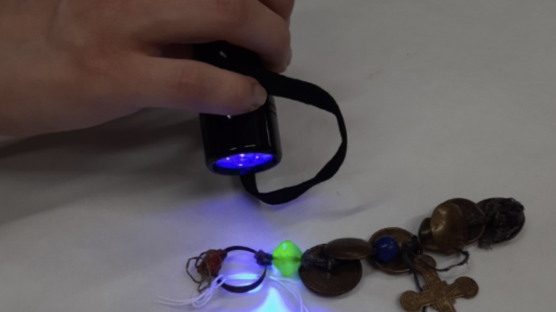 A hand holding a torch shines UV light onto a necklace revealing a bead which glows green.