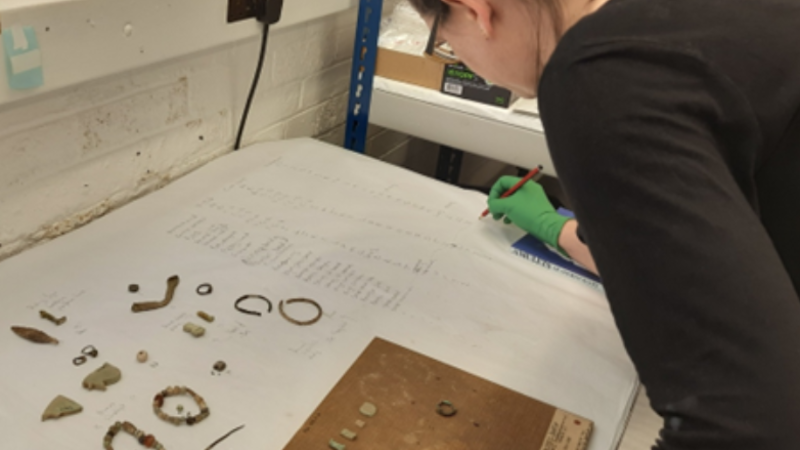 A member of staff works on an old museum display board. Some of the objects removed from the board lie in front of her as she works on a list to identify them all.