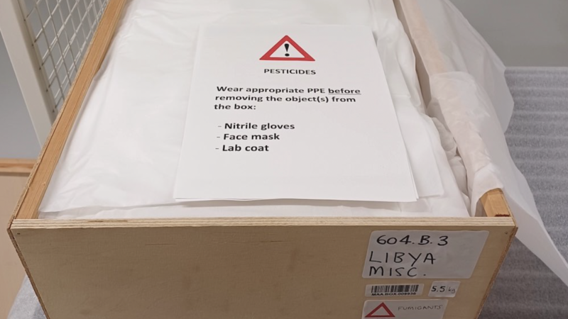An open wooden box, the contents of which is covered with tissue paper. On top lies a notice warning about pesticides and what PPE should be worn and the box is marked with a label alerting to the hazard..