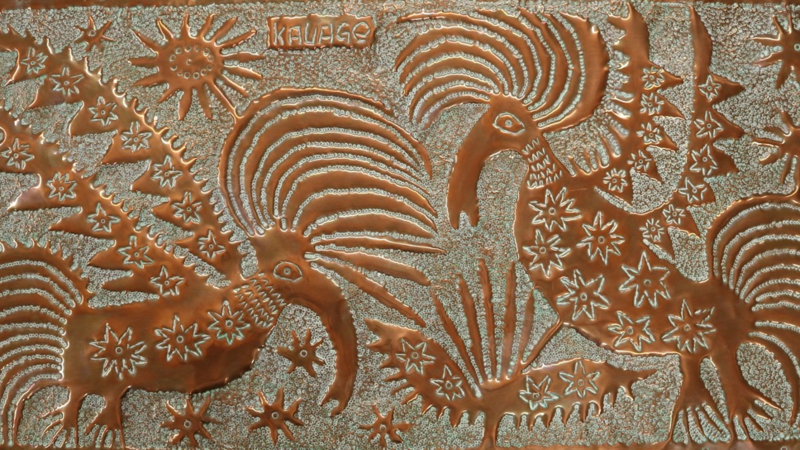 A beaten copper plaque showing two birds of paradise with elaborate plumage in high relief.